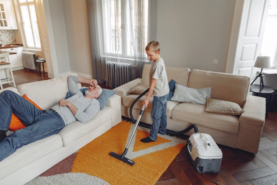 A person kneeling on a patterned area rug in a residential room, using a yellow and black portable vacuum cleaner with a flexible hose to perform surface cleaning. The individual, dressed in casual cl