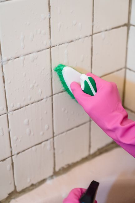 A professional cleaner wearing full protective gear, including a white disposable suit, gloves, a mask, and safety glasses, is engaged in surface cleaning within a kitchen. The cleaner is holding a sp