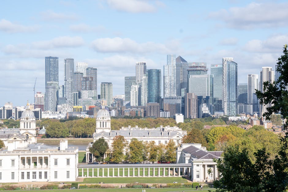 A panoramic view of a large, historic white building with classical architecture, including domed towers and colonnades, situated on a well-maintained grassy park with walking paths and scattered tree
