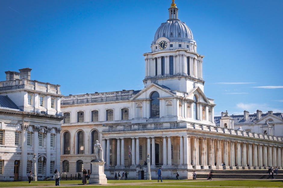 A panoramic view of the Greenwich skyline featuring modern skyscrapers with glass facades reflecting sunlight, set against a bright blue sky with some scattered clouds. In the foreground, a lush green