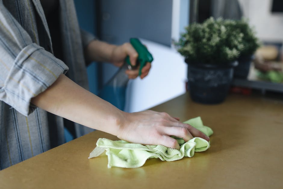 An image of a cleaning cart stationed against a beige tiled wall in a commercial or office setting. The cart contains various cleaning tools and supplies, including a mop with a blue handle, a red bro