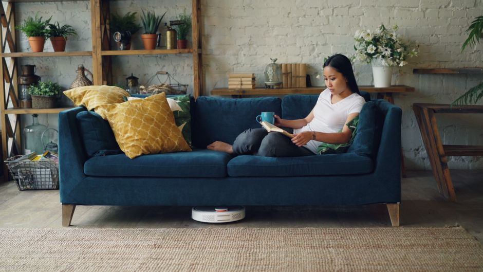A woman sitting on a dark blue fabric sofa in a modern living room, reading a book while holding a blue mug, with a white patterned pillow behind her. The room features wooden shelves with potted plan