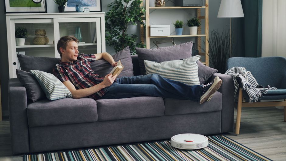 A woman sitting on a dark blue fabric sofa in a modern living room, reading a book while holding a blue mug, with a white patterned pillow behind her. The room features wooden shelves with potted plan